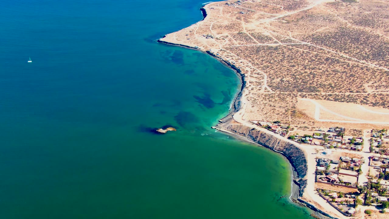 High aerial view of green shallows, rocky coast, desert roads, buildings, and a small white sailboat near the left edge.