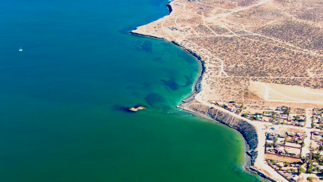 At 0:02, the San Juanico coast remains framed from above, with green water along the rocks, a tiny sailboat offshore, pale desert roads, clustered buildings, and no visible people.