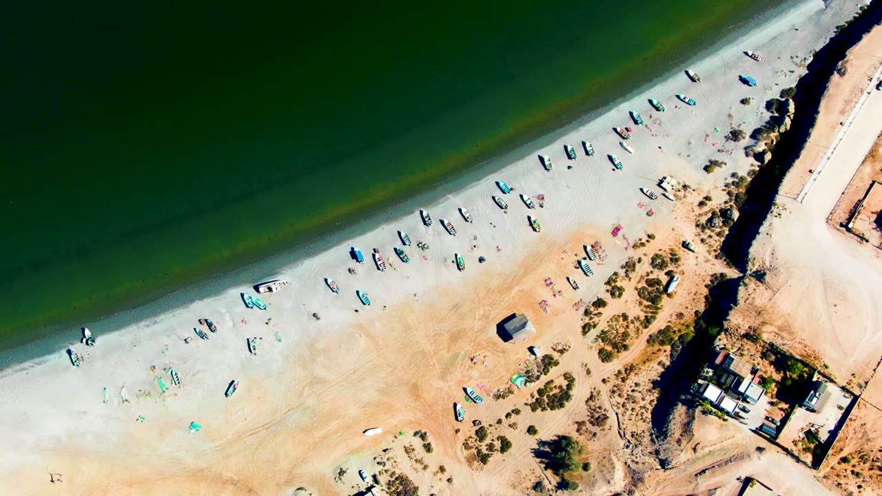 Overhead 0:00 frame of San Juanico beach with green water meeting pale sand, many small boats spread along the shore, structures at the right edge, and no visible people.