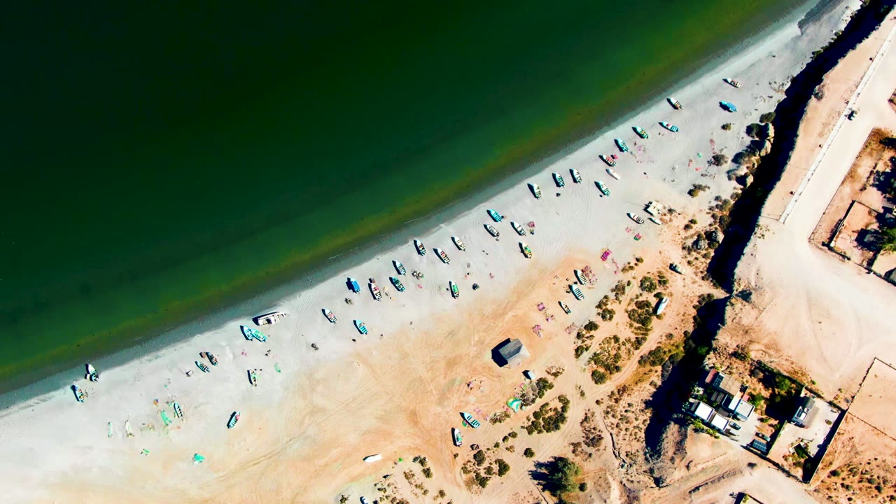 Aerial beach view with green water, clustered small boats, sand, scrub, and right-side buildings.