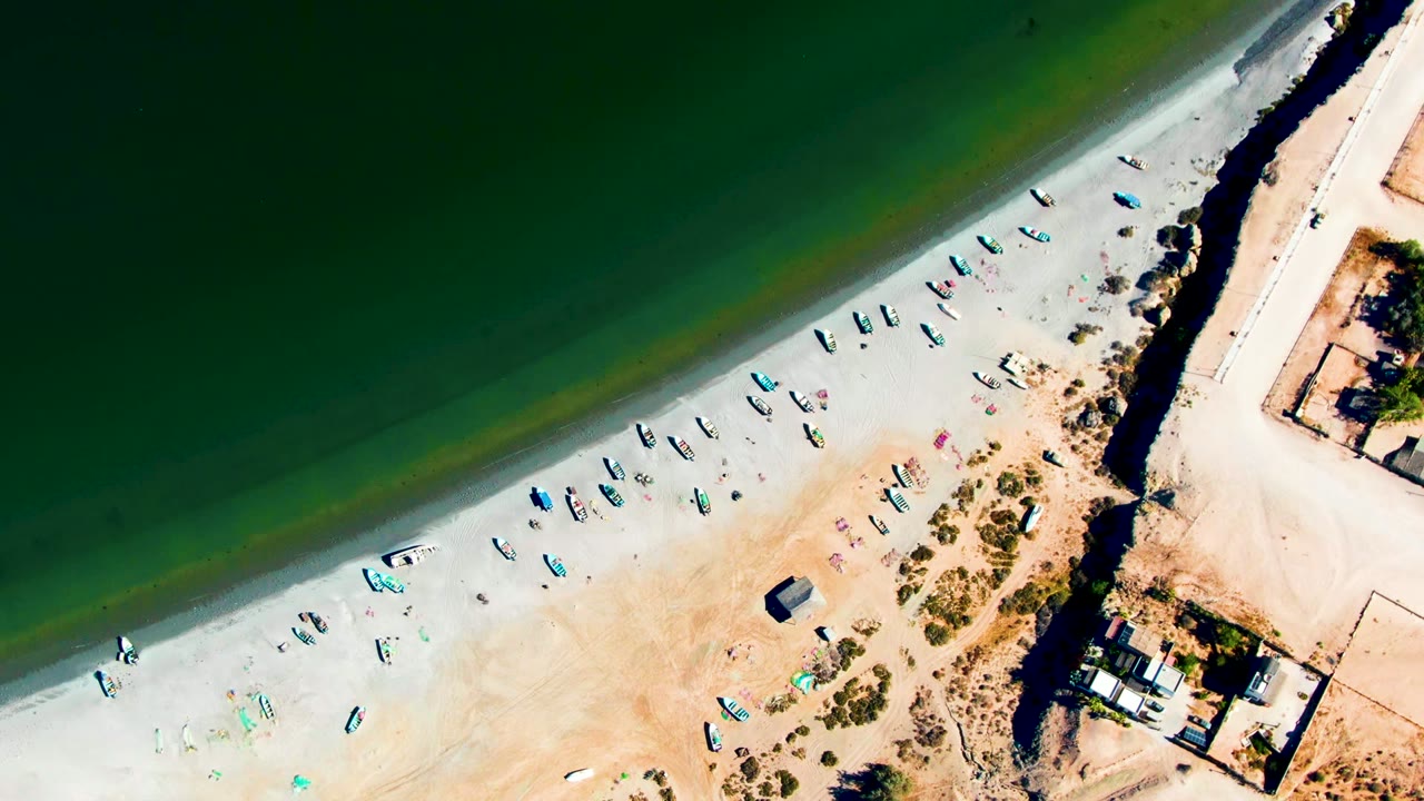 At 0:02, the drone view shows San Juanico's green water across the top, boats scattered on the sandy beach, a straight built edge to the right, and no visible people.
