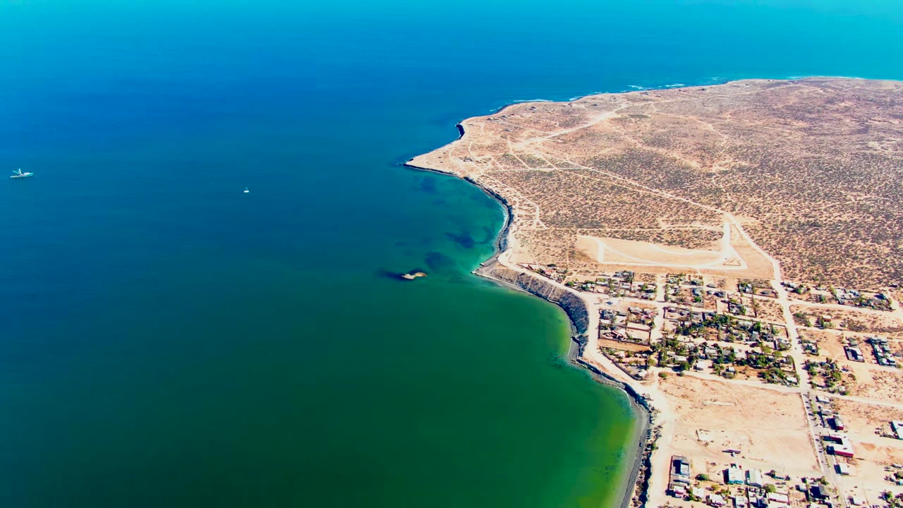 Aerial view over San Juanico, Baja California Sur, with blue-green water on the left, tan arid shoreline, scattered buildings at the lower right, a small white boat offshore, and no visible people.