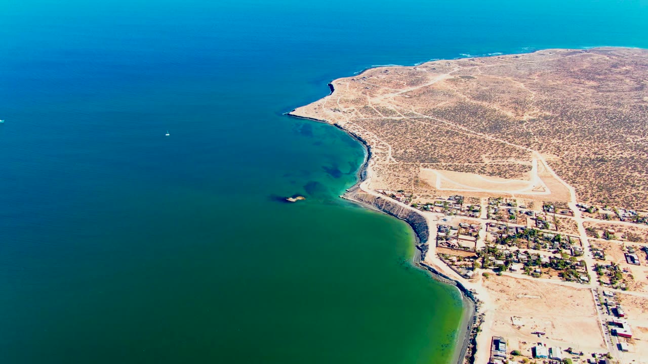 Wide aerial coastline with clear water, desert land, roads, buildings, and a small boat offshore.