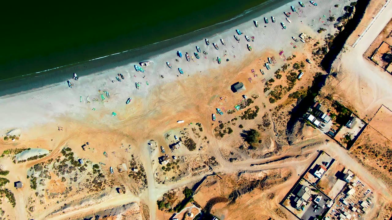 Overhead aerial frame in San Juanico, Baja California Sur, showing rows of small fishing boats on pale sand beside green water, with buildings and dirt tracks at the beach edge.