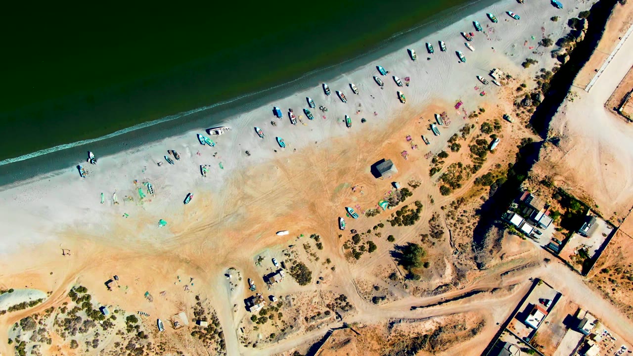 Aerial frame over San Juanico with the green shoreline crossing the top, many small boats on the sand, a dirt road, scrub vegetation, and buildings near the right edge.