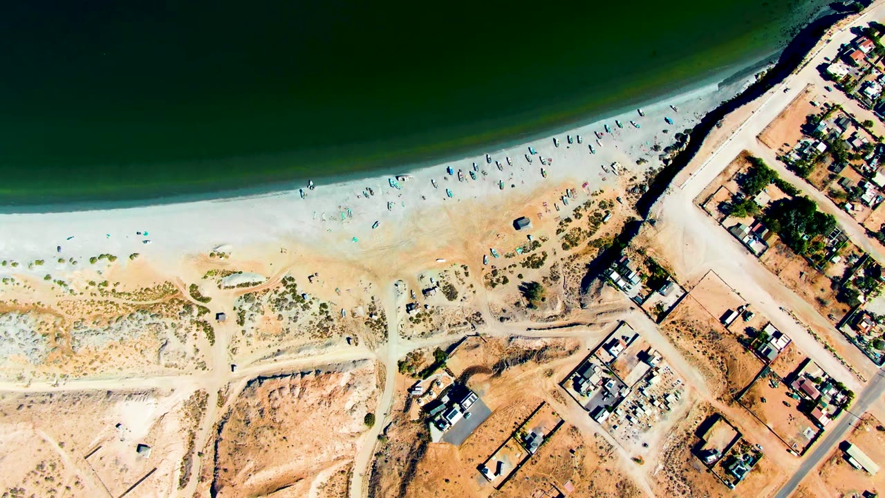 High aerial frame over San Juanico, Baja California Sur, with dark green ocean at the top, pale beach through the center, tiny beachgoers, and houses along dirt roads at right.