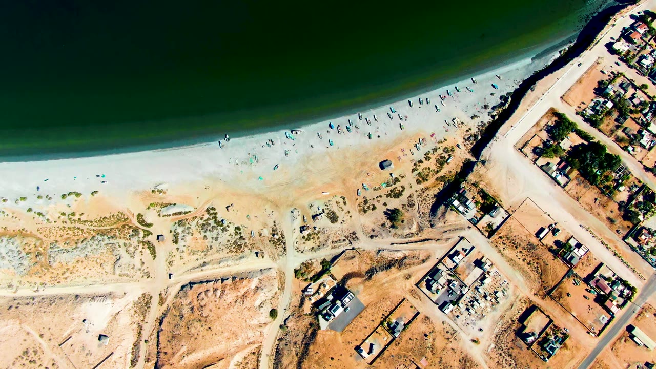 Aerial beach frame with ocean, people on sand, roads, and houses.