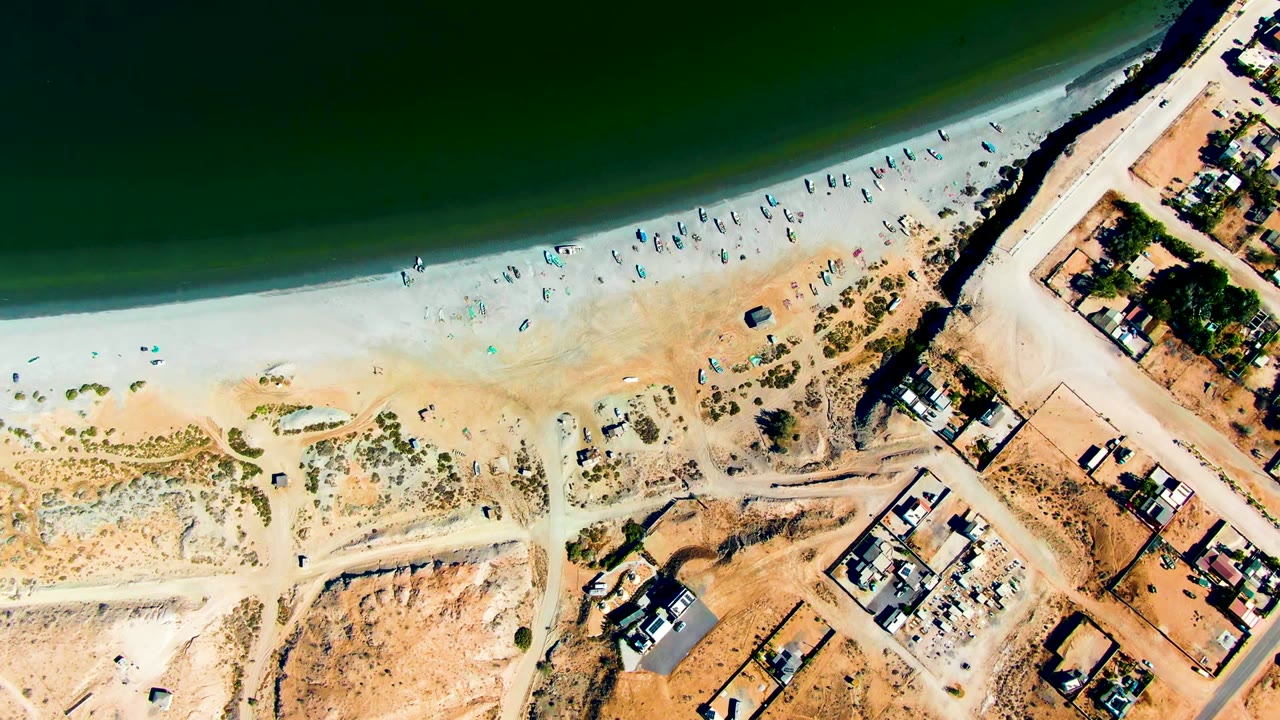Aerial frame over San Juanico showing a broad pale sand band, scattered beachgoers near the water, tan undeveloped ground, and a cluster of residential buildings at lower right.