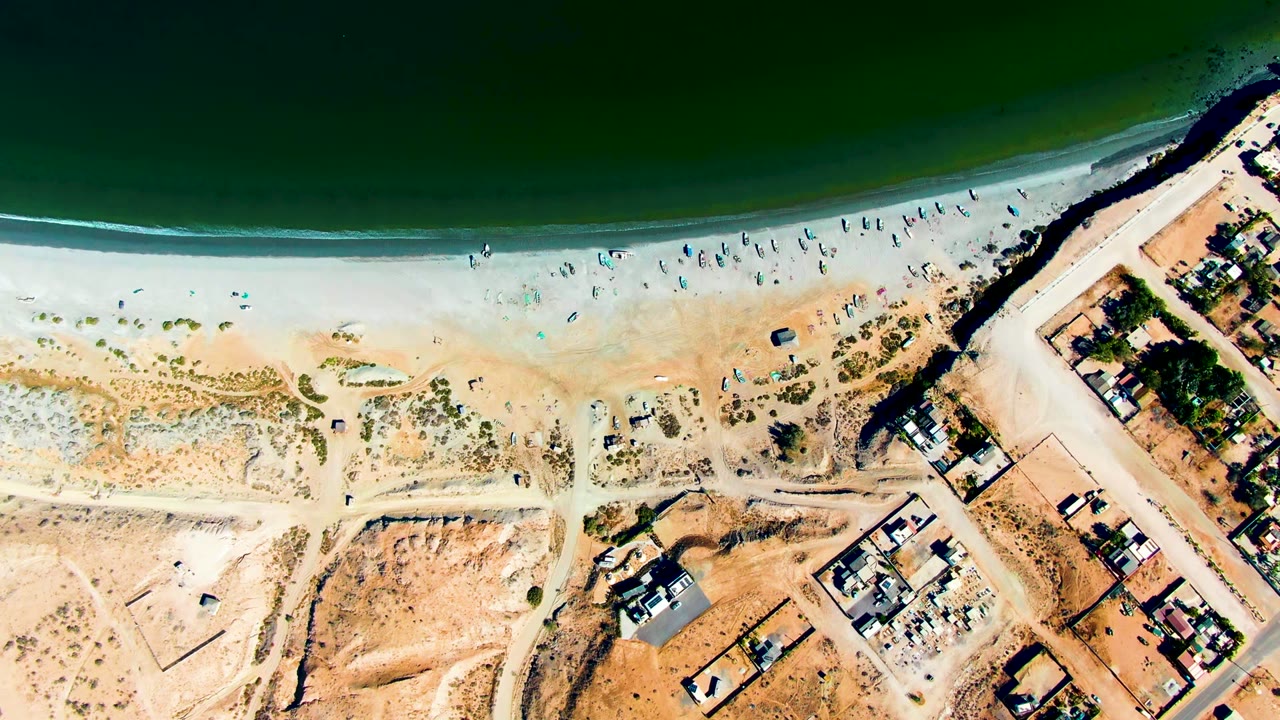 Top-down aerial frame of San Juanico, Baja California Sur with dark green ocean above a pale sandy beach, scattered beachgoers, dry terrain, roads, and houses along the right edge.