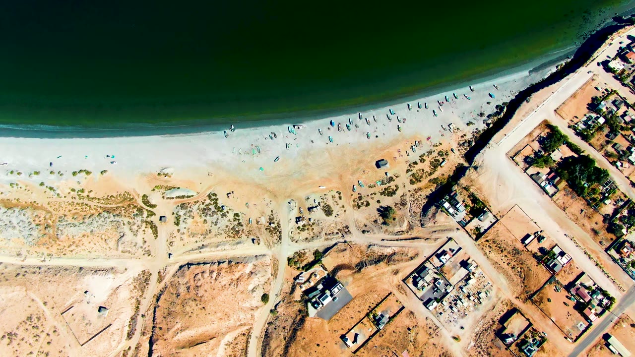 Aerial frame of San Juanico beach with green water at the top, a pale surf edge, fewer small figures along the sand, and a diagonal cluster of houses and roads to the right.
