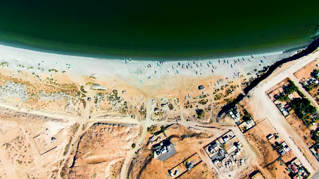 Top-down aerial frame of San Juanico, Baja California Sur, with dark green ocean above a pale sandy beach, scattered beachgoers, dune paths, a road, and coastal homes.