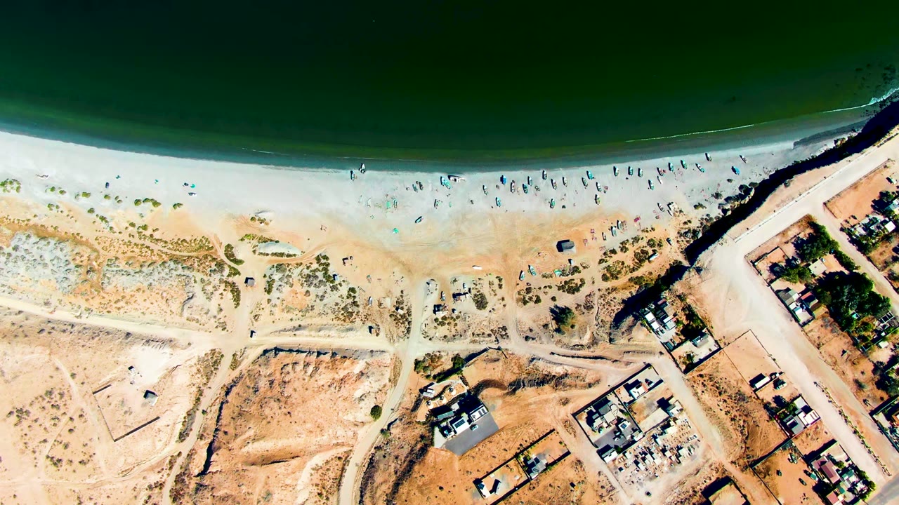 Top-down San Juanico beach frame with gentle surf at the top, small beachgoers spread along the sand, dune scrub, a diagonal road, and clustered coastal homes.