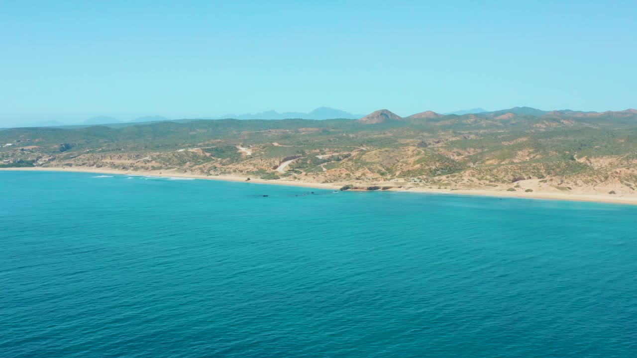 Aerial view at El Tule Los Cabos showing blue ocean in the foreground, sandy shoreline and dry hills beyond, a small dark boat offshore, and no visible people.