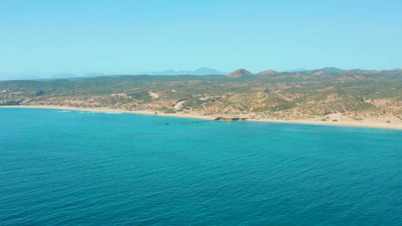 Wide aerial coastline with turquoise water, pale beach, hills, and a small boat near shore.