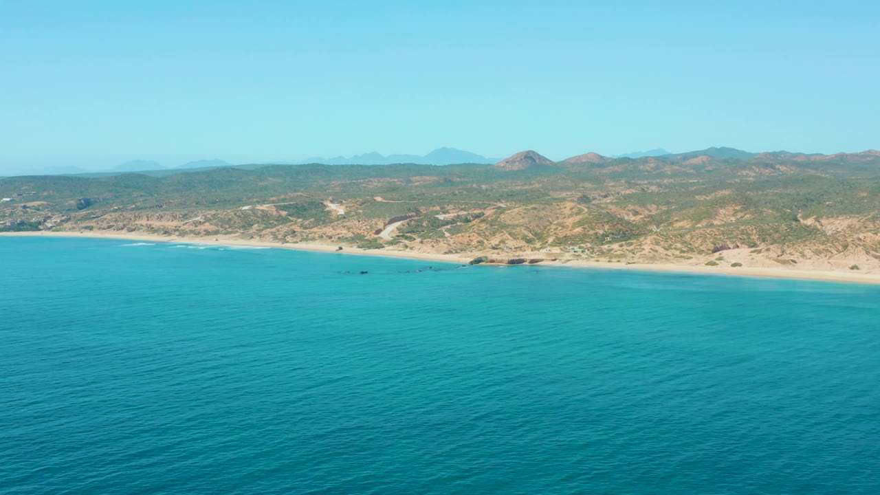 Aerial frame at El Tule Los Cabos showing broad blue water, a sandy beach line, scrub-covered terrain, distant low mountains, a small dark boat near the shore, and no visible people.