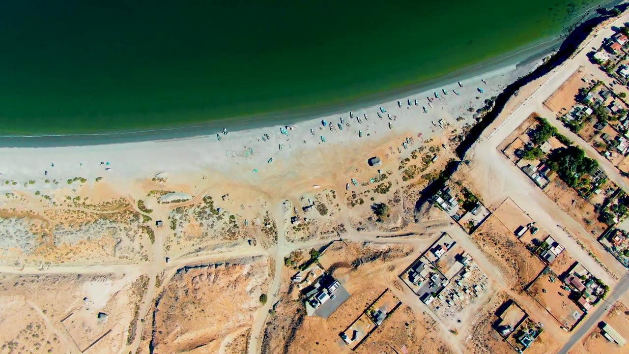 Overhead view in San Juanico, Baja California Sur, Mexico showing green ocean water, a pale sandy beach with scattered people, dirt tracks, and coastal buildings at the start of the clip.