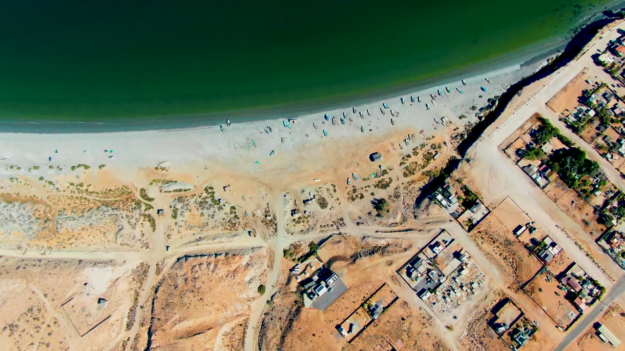 Aerial shoreline with beachgoers, green water, sand lots, roads, and buildings.