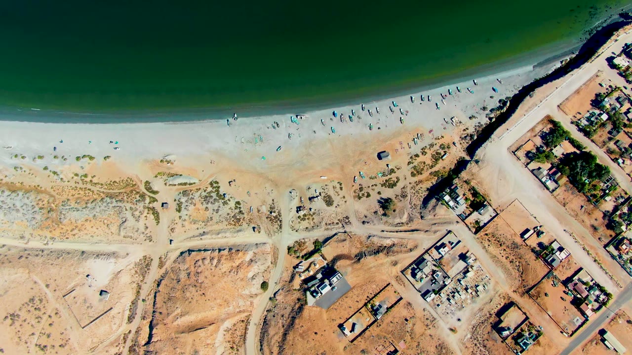 Mid-clip overhead frame in San Juanico showing the green ocean band, a pale beach with fewer visible beachgoers than the start, and construction lots and buildings beside dirt roads.