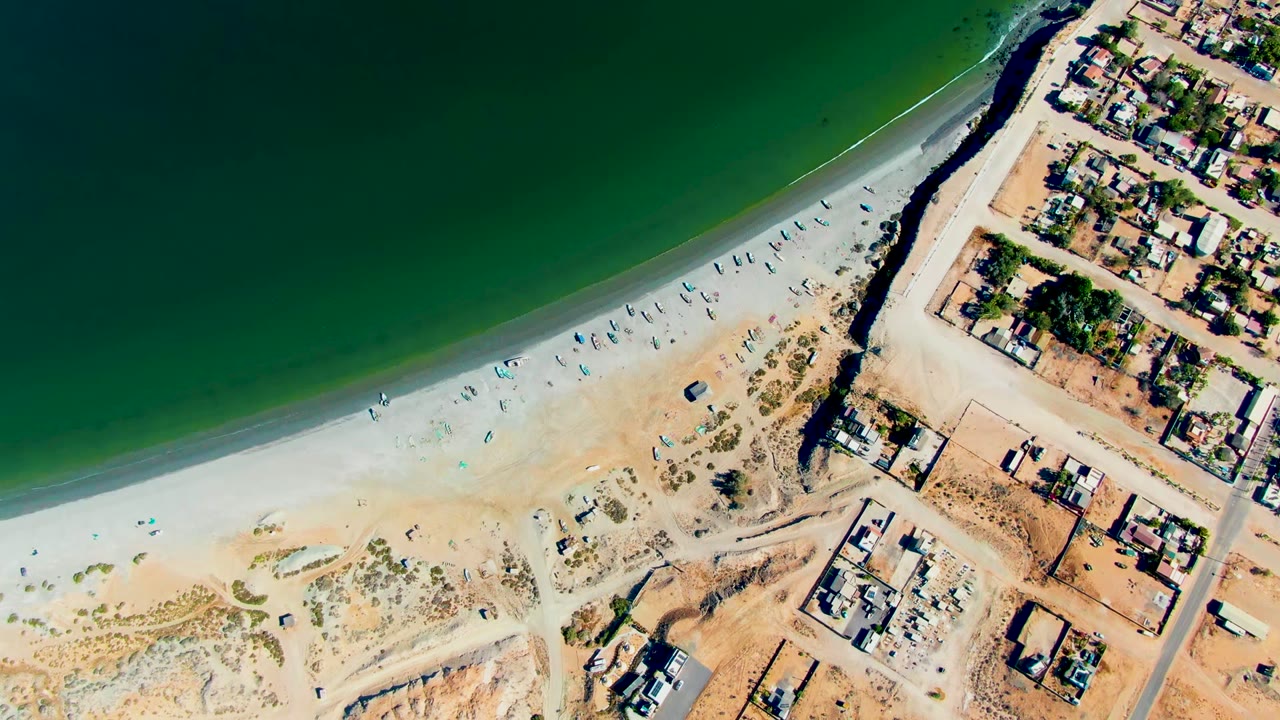 Overhead view of San Juanico beach with green ocean at upper left, pale sand with umbrellas and tiny people, dry land below, and houses along the right edge.