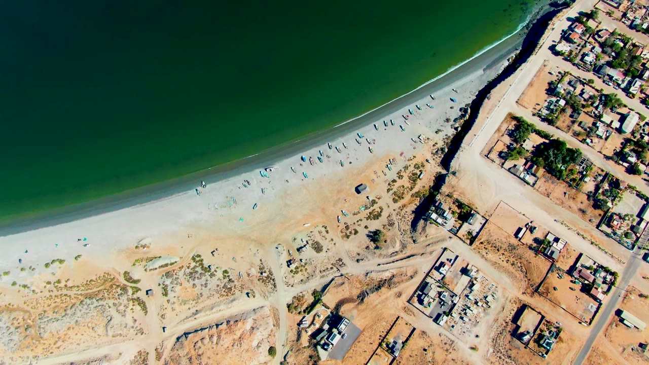 Top-down view of San Juanico beach where green water curves along the sand, umbrellas dot the shore, small people are visible, and homes line the right side.