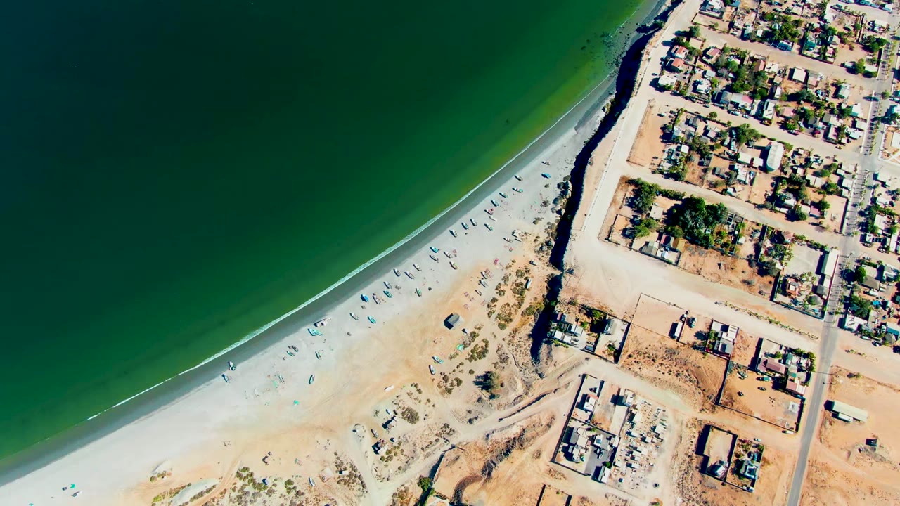Overhead aerial frame of San Juanico, Baja California Sur with green ocean water curving along pale sand, low coastal buildings to the right, and no visible people.