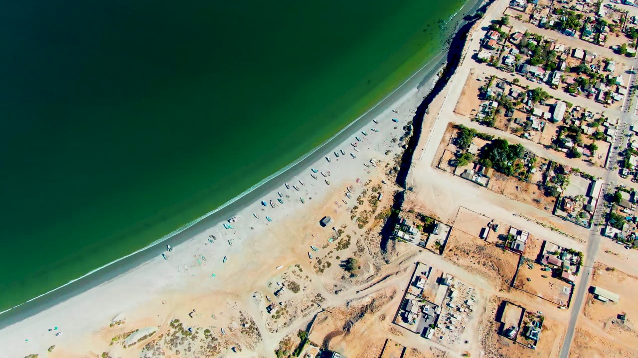 Aerial view over San Juanico with the green ocean filling the upper frame, a bright sandy shoreline below it, beachfront roads, buildings, and no visible people.