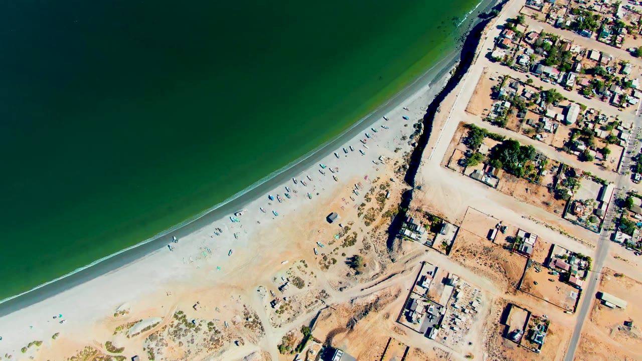 Top-down coastal frame in San Juanico showing a thin surf line, open sand with scattered objects, dense low buildings at the right edge, and no visible people.