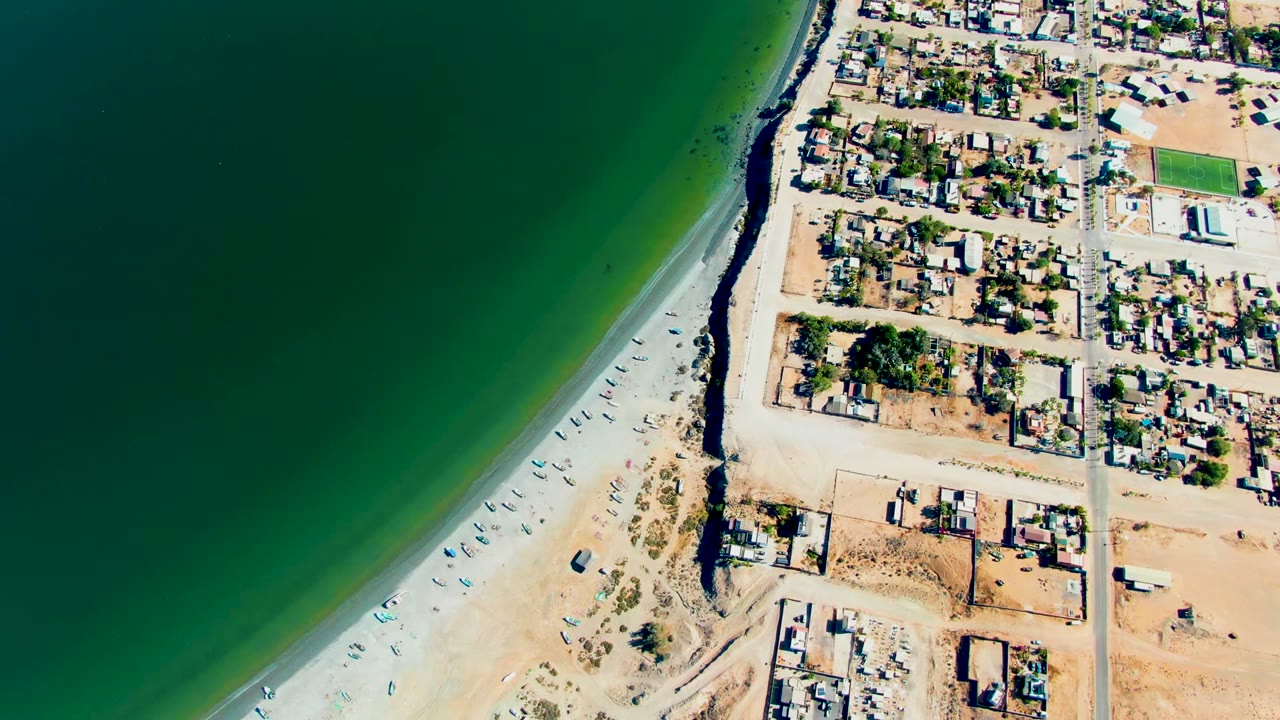Overhead view of San Juanico, Baja California Sur, with green water on the left, pale sand and small boats along the shore, and residential lots on the right, with no visible people.