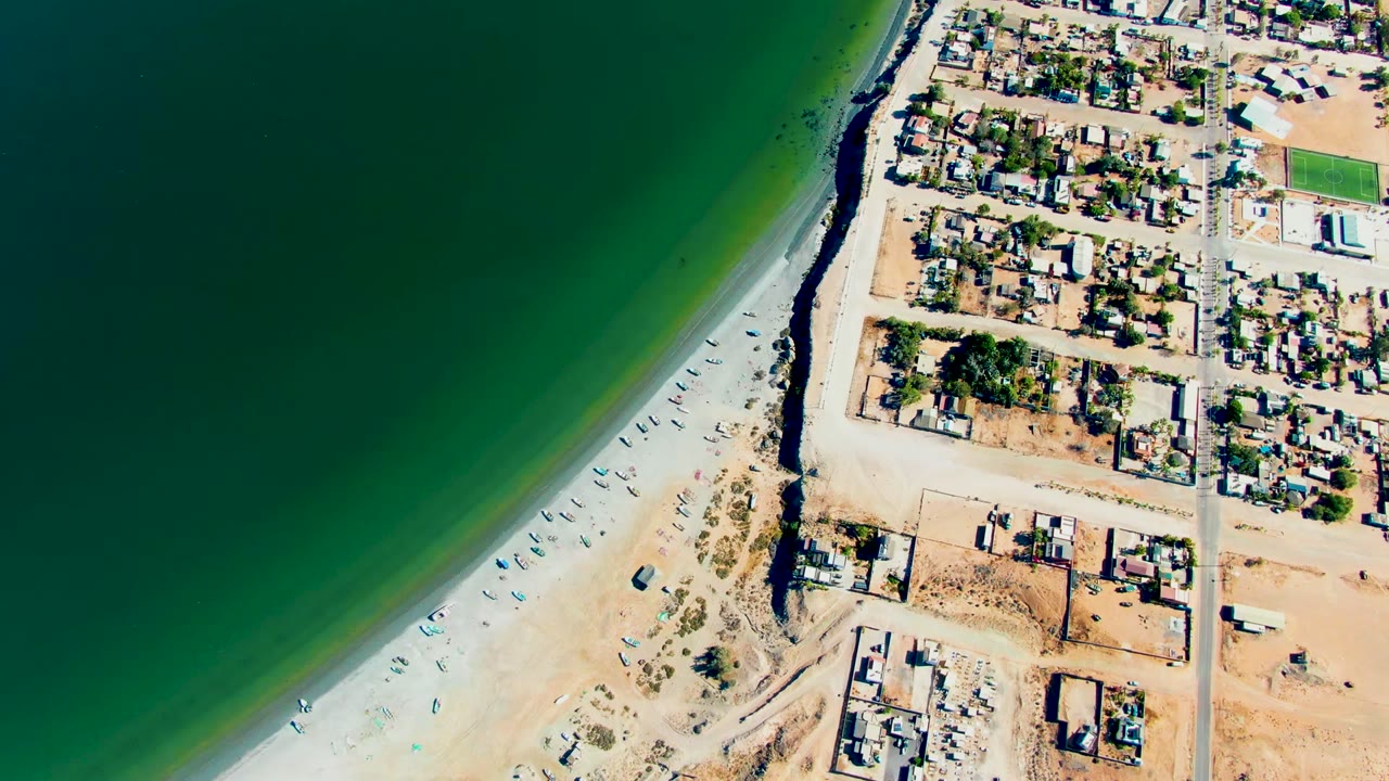 High aerial frame over San Juanico showing the green water band, curved pale beach with small vessels, and the residential area pressed close to the shore, with no visible people.