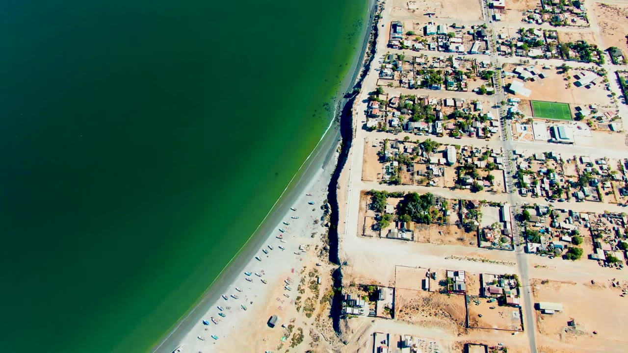 Aerial frame over San Juanico, Baja California Sur, with green ocean on the left, a pale beach and road through the middle, low homes on the right, and no visible people.