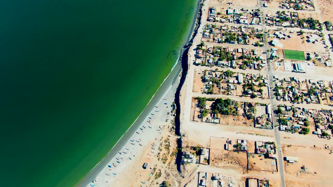 Overhead coastal view with beach road, small vehicles, homes, and green water.