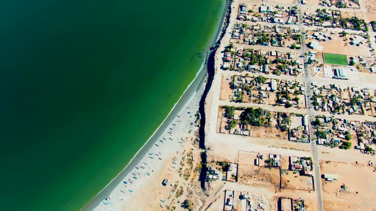 High aerial frame in San Juanico with the road tracing the beach edge, green ocean water to the left, neighborhood blocks to the right, and no visible people.