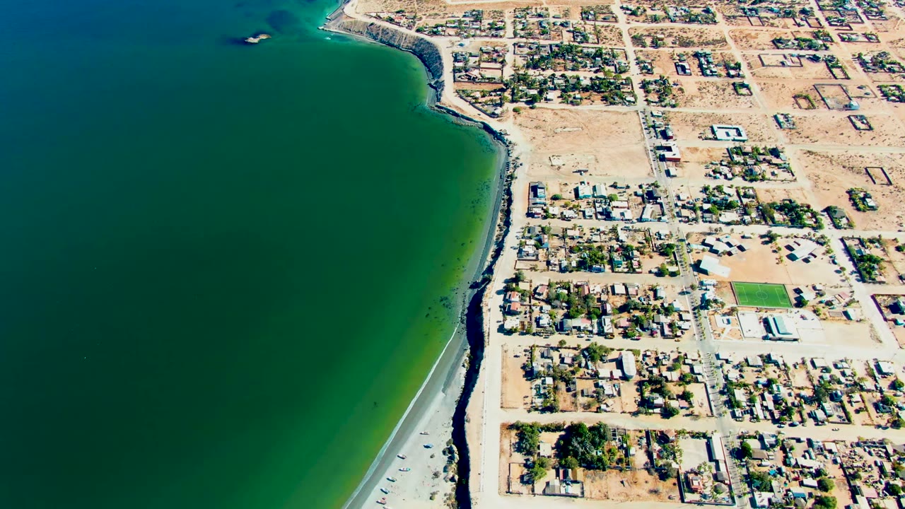 Opening aerial frame over San Juanico, Baja California Sur, with green-blue ocean, a small offshore boat near the upper shoreline, sandy beach, residential lots, and no visible people.