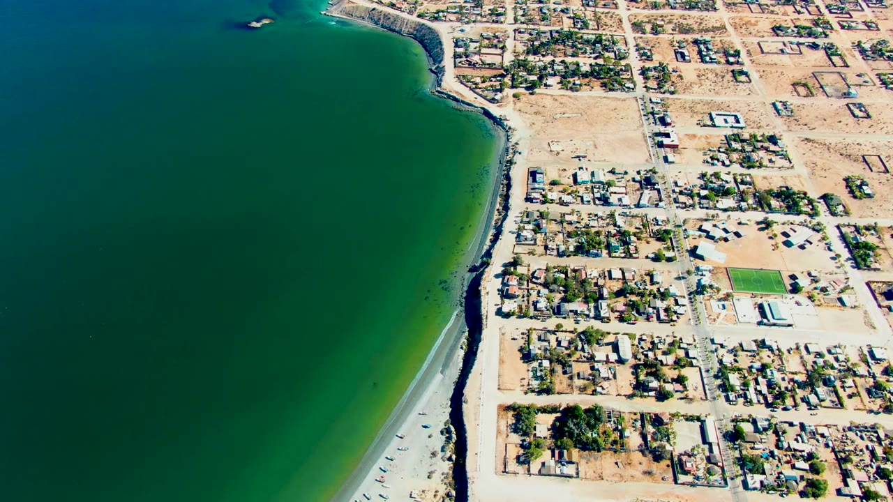 San Juanico aerial still with the ocean filling the left side, sandy beach and rock line dividing the frame, residential buildings to the right, and no visible people.