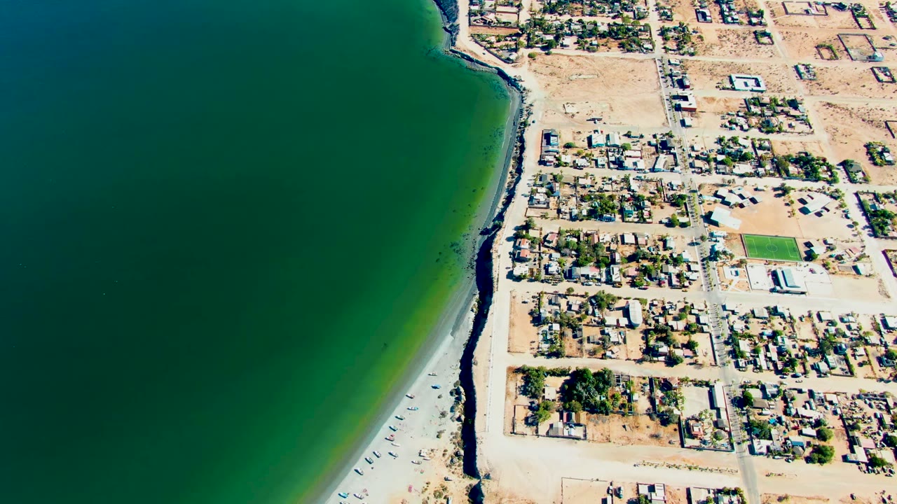 Aerial frame over San Juanico, Baja California Sur, Mexico with dark green ocean on the left, a narrow sandy shoreline, coastal houses and roads on the right, and no visible people.