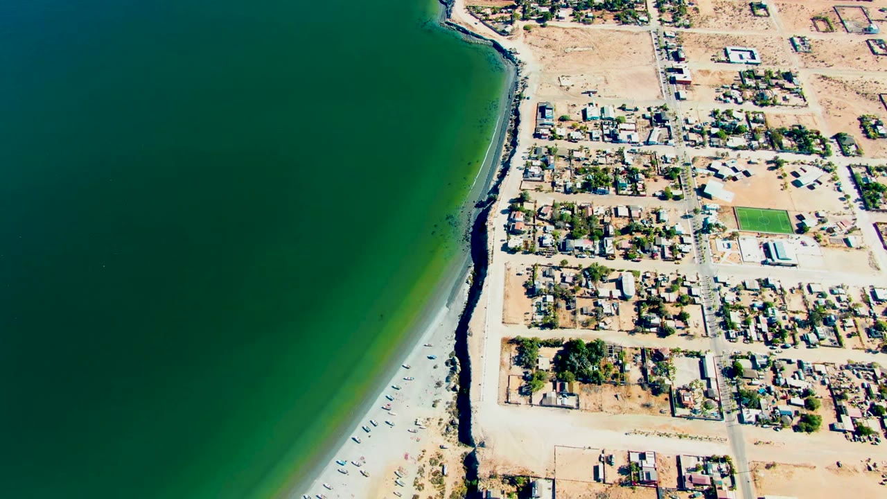 High aerial frame in San Juanico with the ocean occupying most of the left side, a curved sandy shore below, residential buildings and streets to the right, and no visible people.