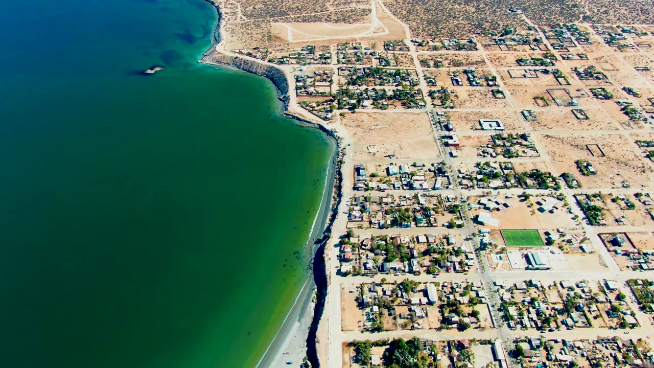 High aerial view of San Juanico, Baja California Sur, with green ocean water on the left, a sandy beach edge, roads, and low homes inland, with no visible people.