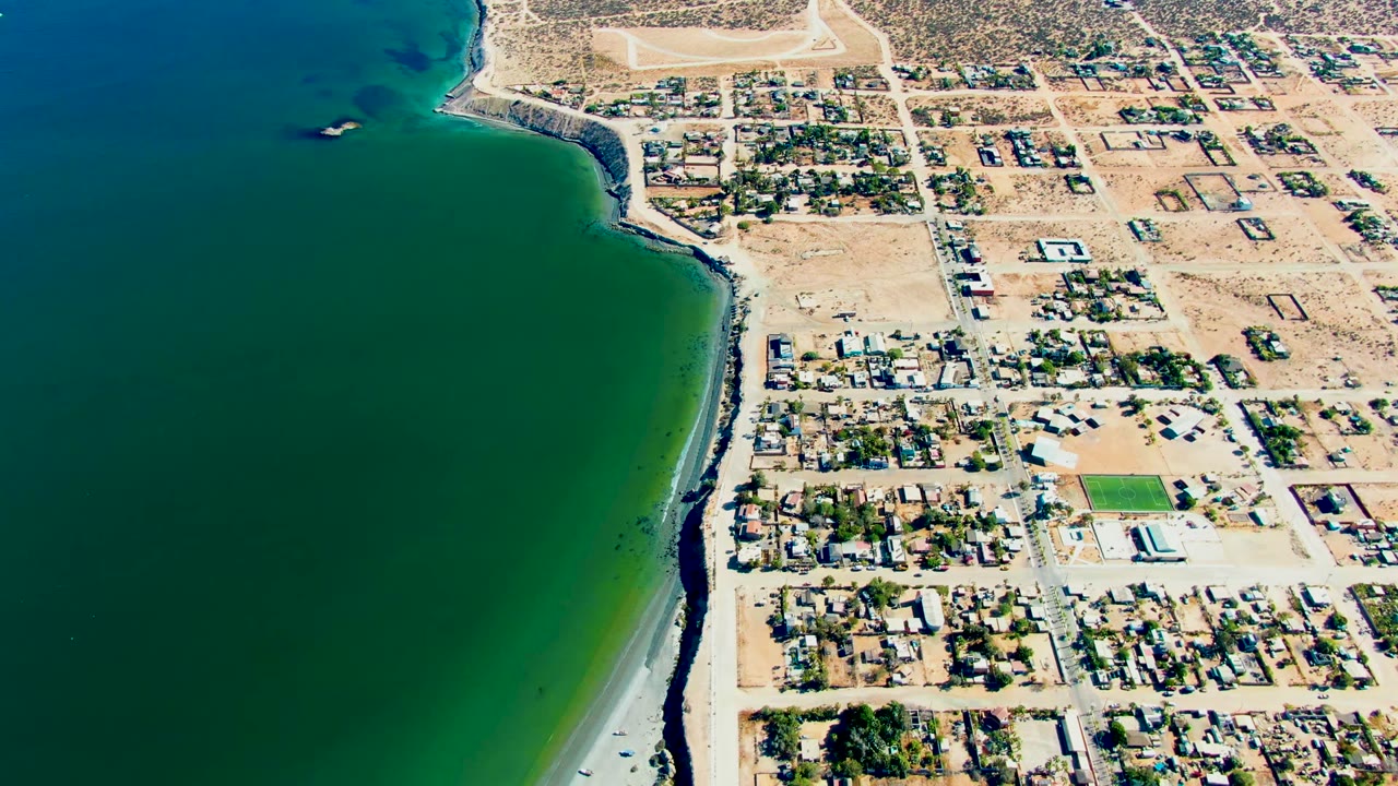 San Juanico coastline seen from above, with deep green water, pale sand, a road running near the beach, and low houses set among dry lots, with no visible people.