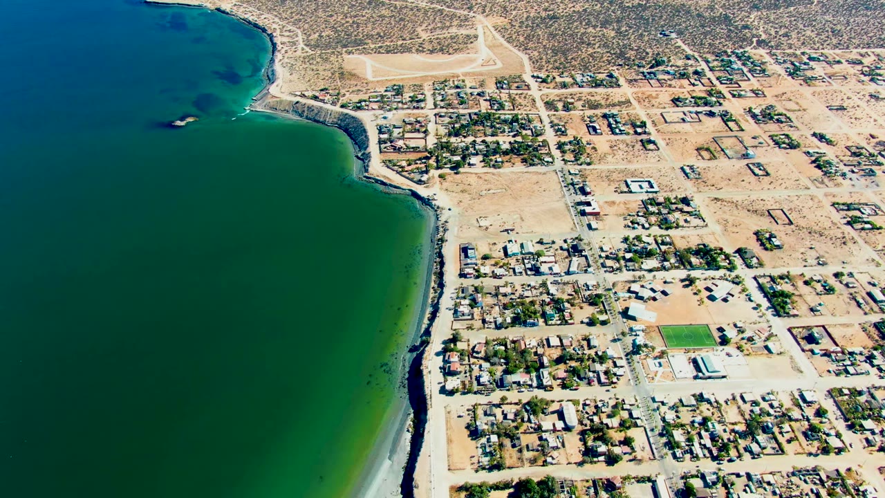 Aerial view over San Juanico, Baja California Sur, with green bay water on the left, a pale beach edge, dry streets, houses, and no visible people.