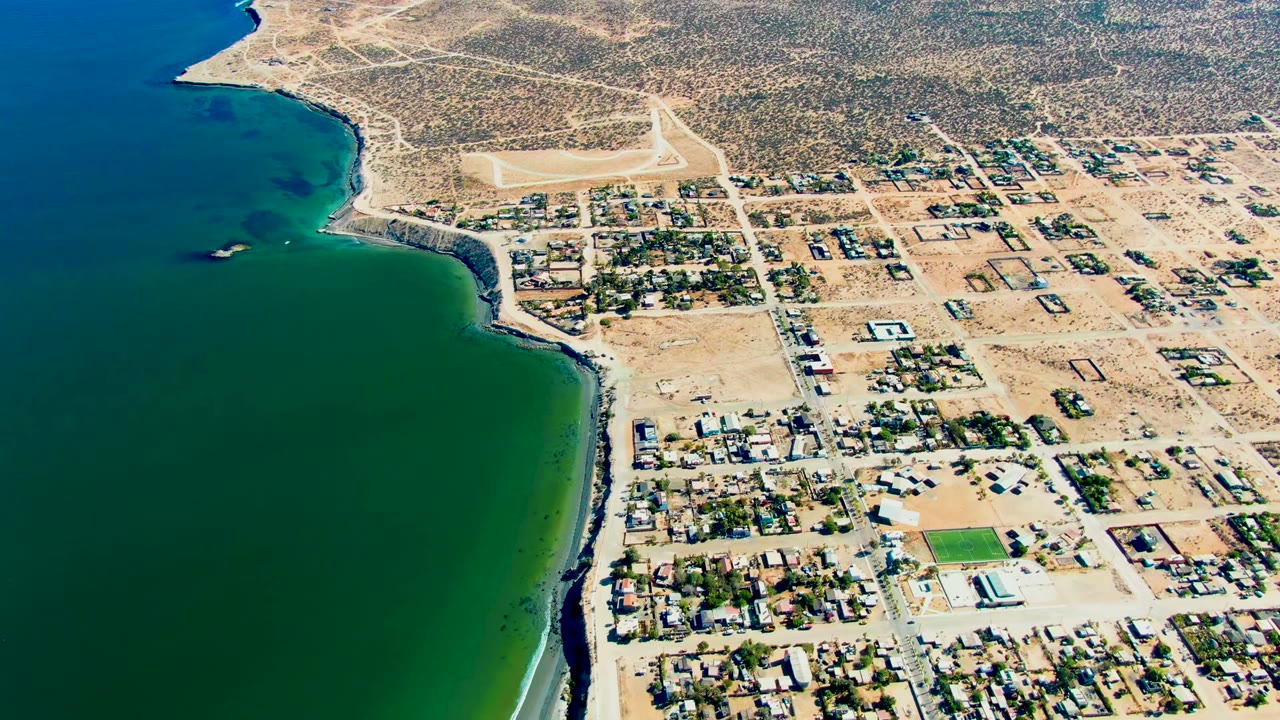 Aerial frame over San Juanico, Baja California Sur showing blue-green water on the left, a curved beach, grid streets, houses, dry terrain, and no visible people.