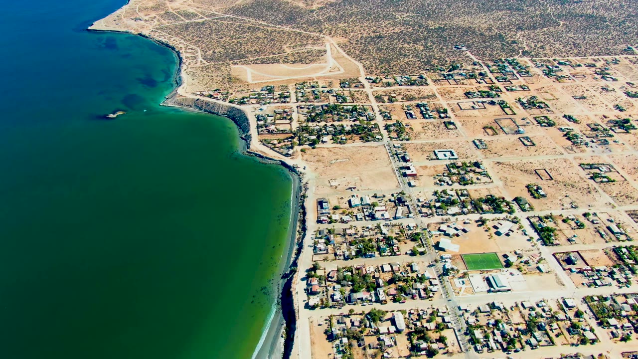 High aerial frame of San Juanico showing calm blue-green water, a pale beach strip, rectangular street blocks, scattered homes, dry background terrain, and no visible people.