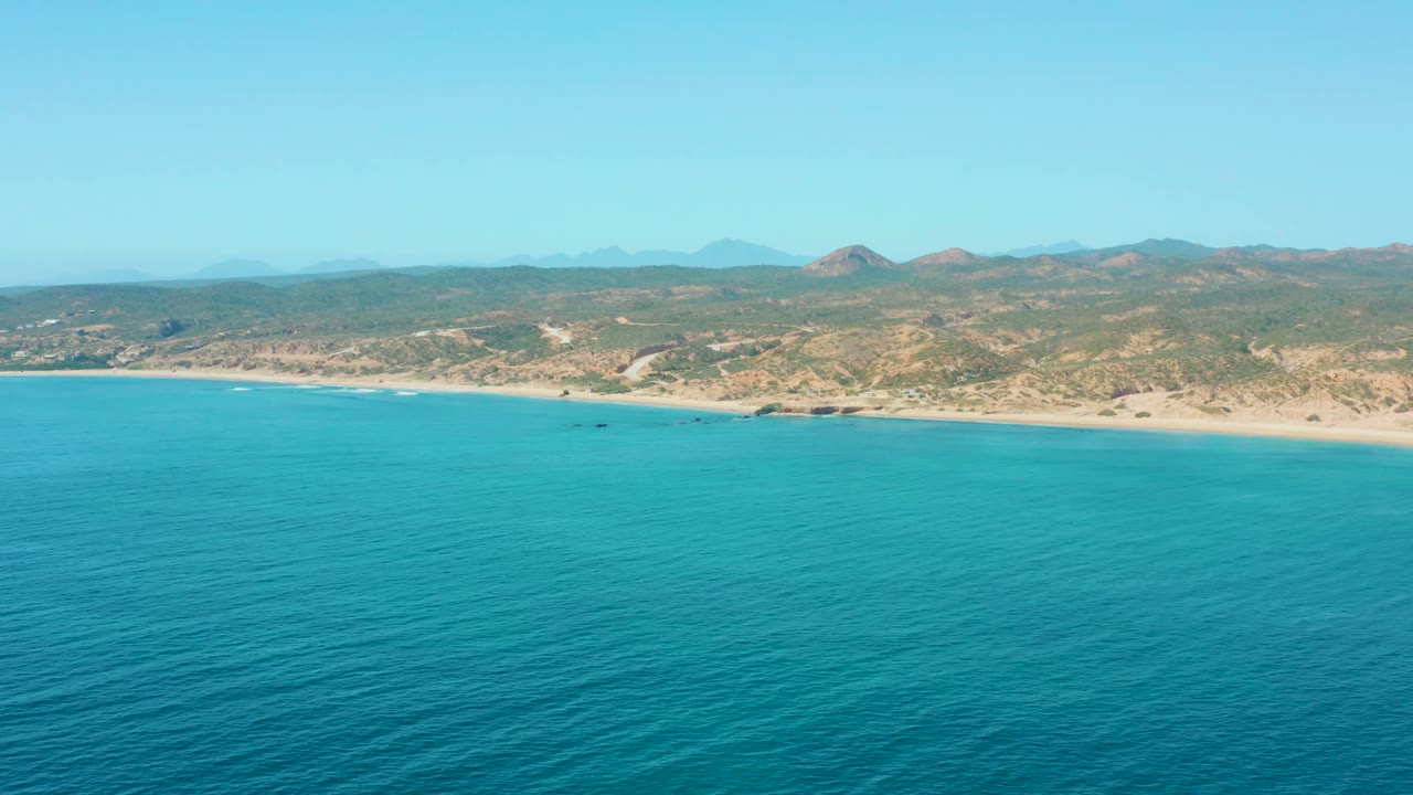 Opening frame of El Tule Los Cabos with turquoise ocean in the foreground, a sandy beach beyond it, low hills in the distance, and no visible people.