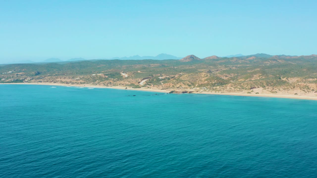 Aerial coastline view with open blue water and pale beach below distant hills.