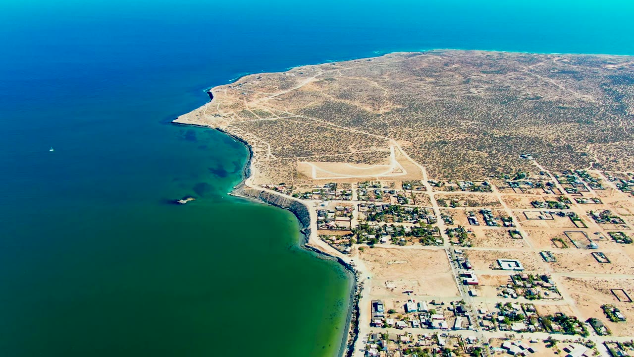 Aerial view over San Juanico, Baja California Sur, Mexico, with blue-green ocean at left, a sandy shoreline, town buildings, an airstrip, dry scrubland, and no visible people.