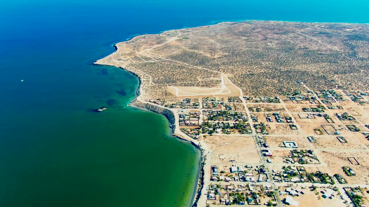 A high aerial view of San Juanico with the coastline sliding along the left side, the airstrip centered inland, rectangular town parcels, open desert, and no visible people.