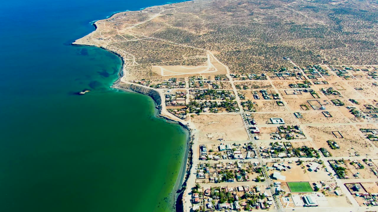 Opening aerial frame over San Juanico, Baja California Sur, with green ocean on the left, a curving shoreline, dry lots, homes, dirt roads, arid hills, and no visible people.