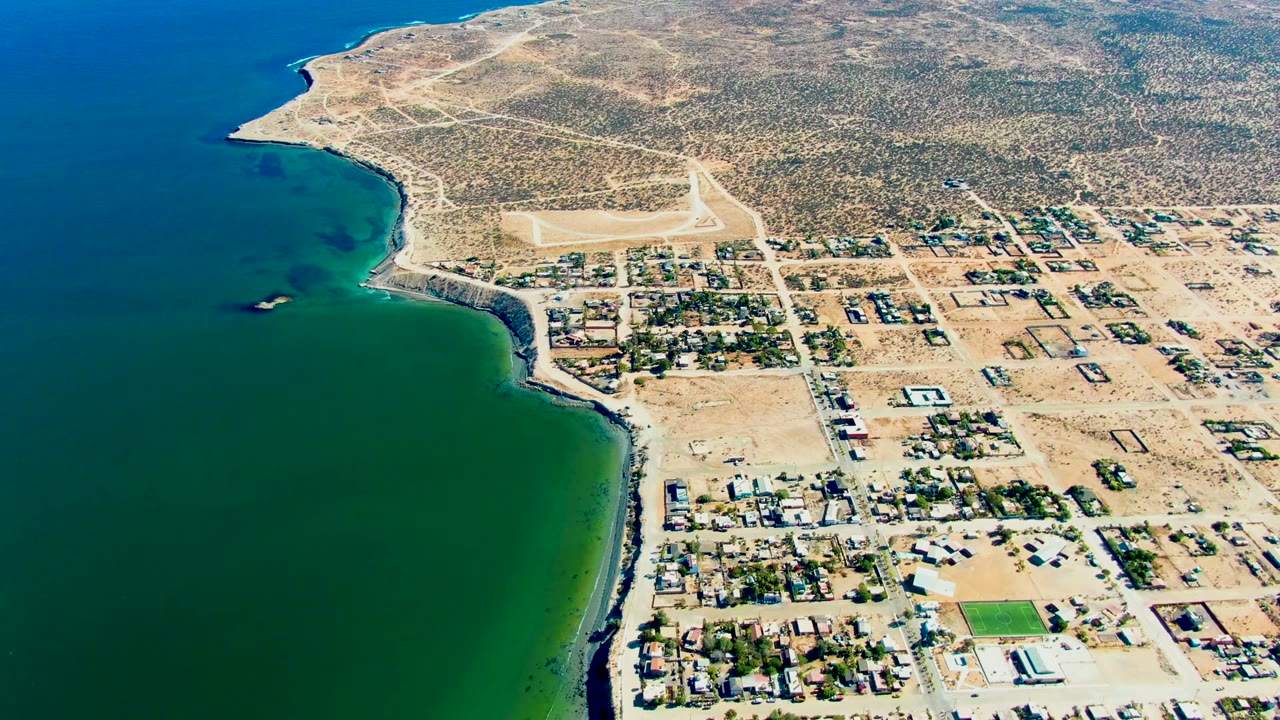Drone view of a curved shoreline, ocean water, houses, roads, and desert hills.