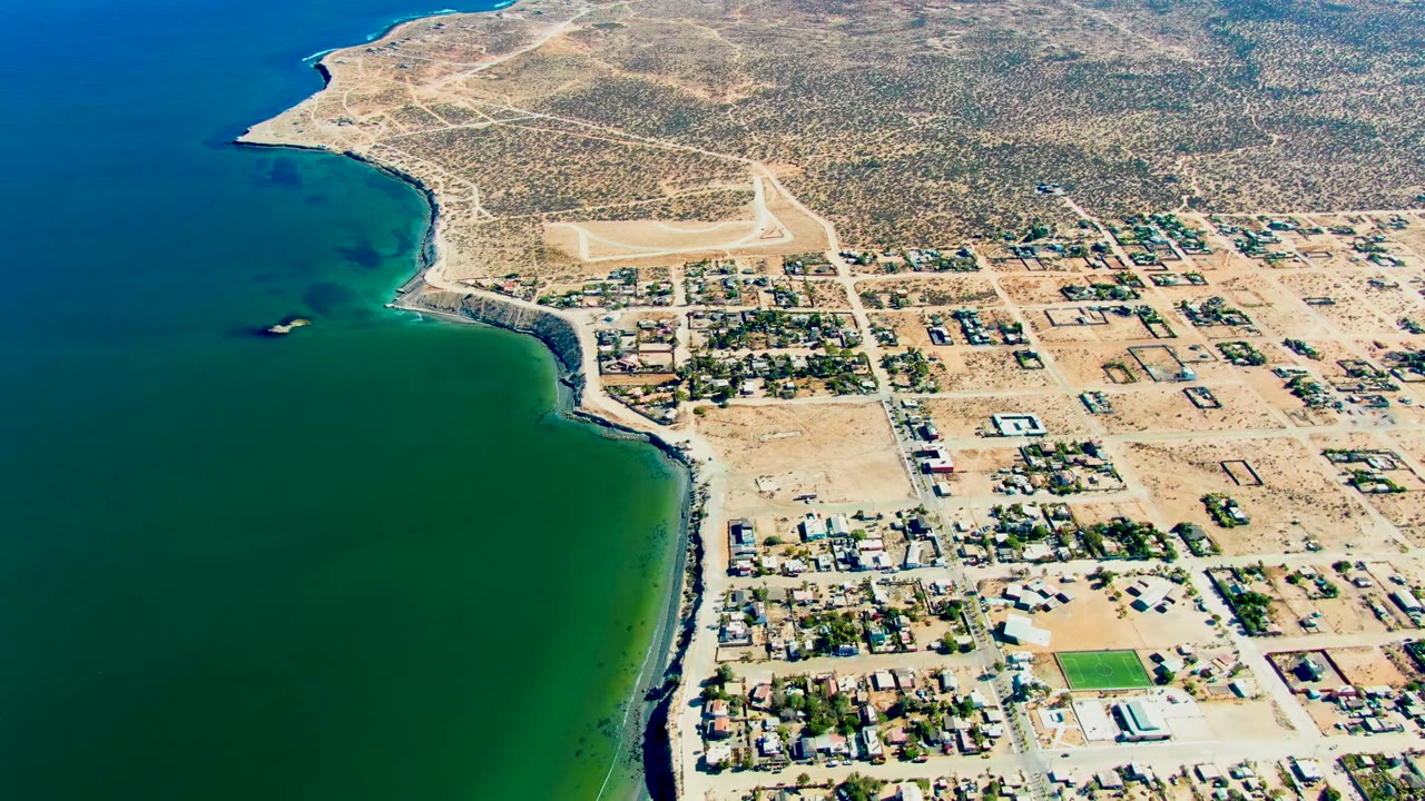 At 0:02, the drone frame shows San Juanico's beach edge, darker green water near shore, scattered homes and rectangular lots, pale roads, arid hills, and no visible people.
