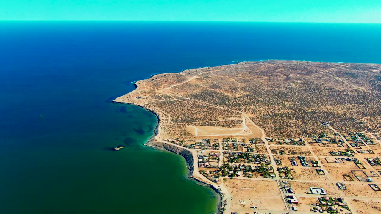 Opening aerial frame over San Juanico, Baja California Sur, showing green coastal water, deep blue ocean, a dry shoreline settlement, a pale runway inland, and no visible people.