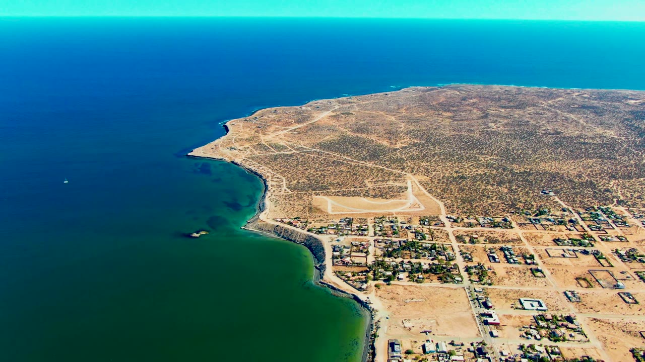 Mid-clip aerial view of San Juanico showing the runway more centrally against tan desert ground, with town structures near the shore and no visible people.